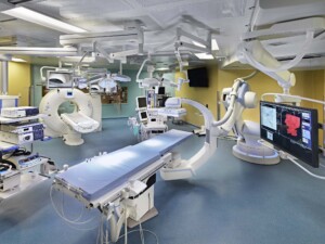 The TRIGOR-A operating room at Toronto General Hospital, featuring a Siemens CT scanner, an angiography machine, an operating table, ceiling-mounted surgical lights, and multiple monitors displaying medical imaging.