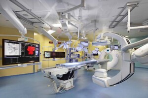 The TRIGOR-A operating room at Toronto General Hospital, featuring a Siemens Flash-CT scanner, an Artis Zeego C-arm, an operating table, ceiling-mounted surgical lights, and multiple monitors displaying medical imaging.