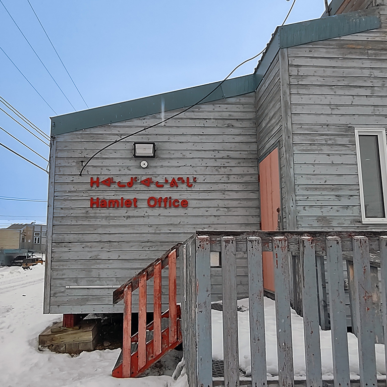 Snowy front corner of Grise Fiord Hamlet Office; red syllabic and English sign on grey timber wall beside weathered ramp.