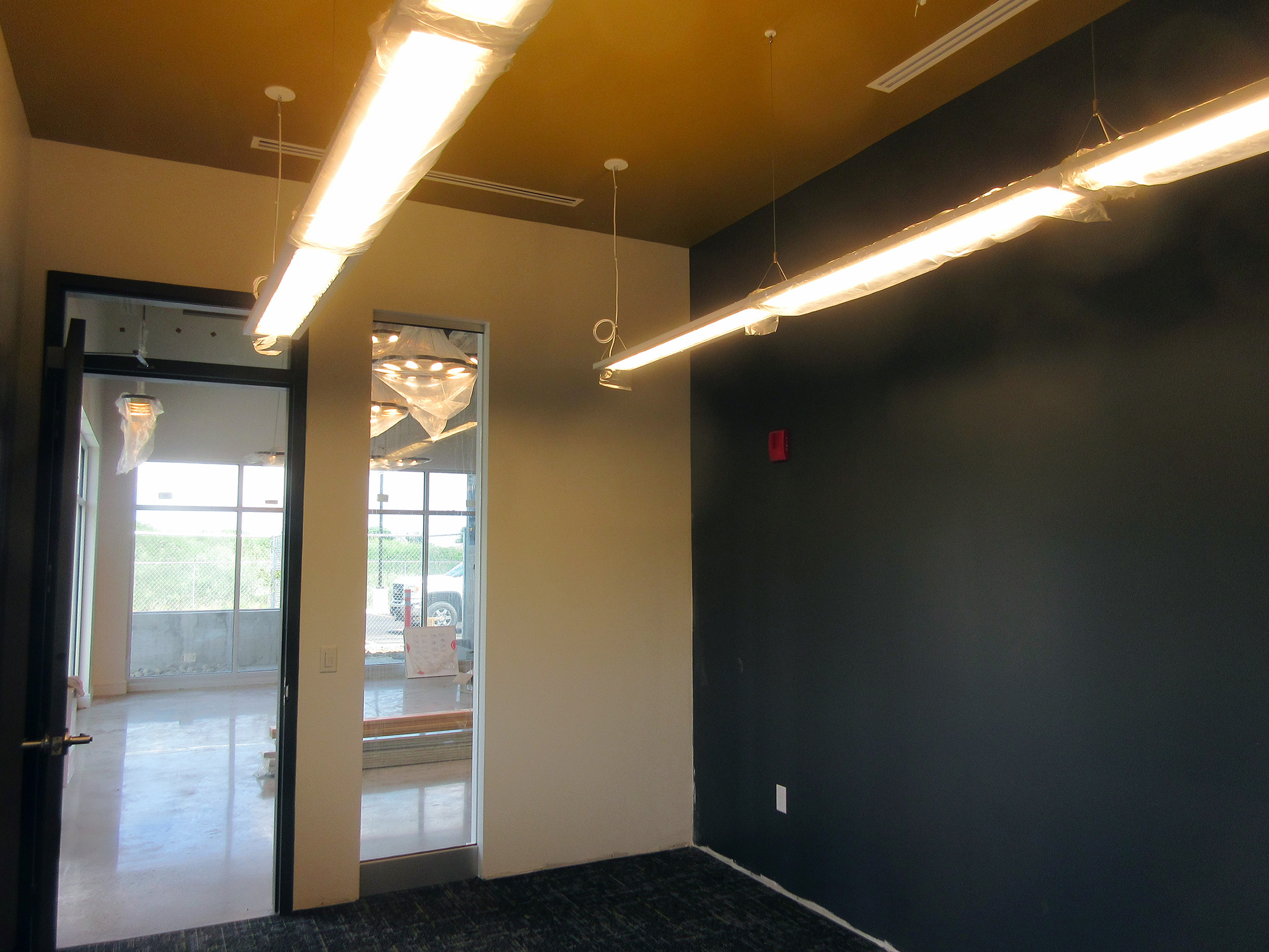 Latitude Air Ambulance 11 Small office at the Latitude Air Ambulance facility with a dark feature wall, gold ceiling, and suspended linear LED lights still wrapped in protective plastic; a glass doorway opens to the lobby beyond.
