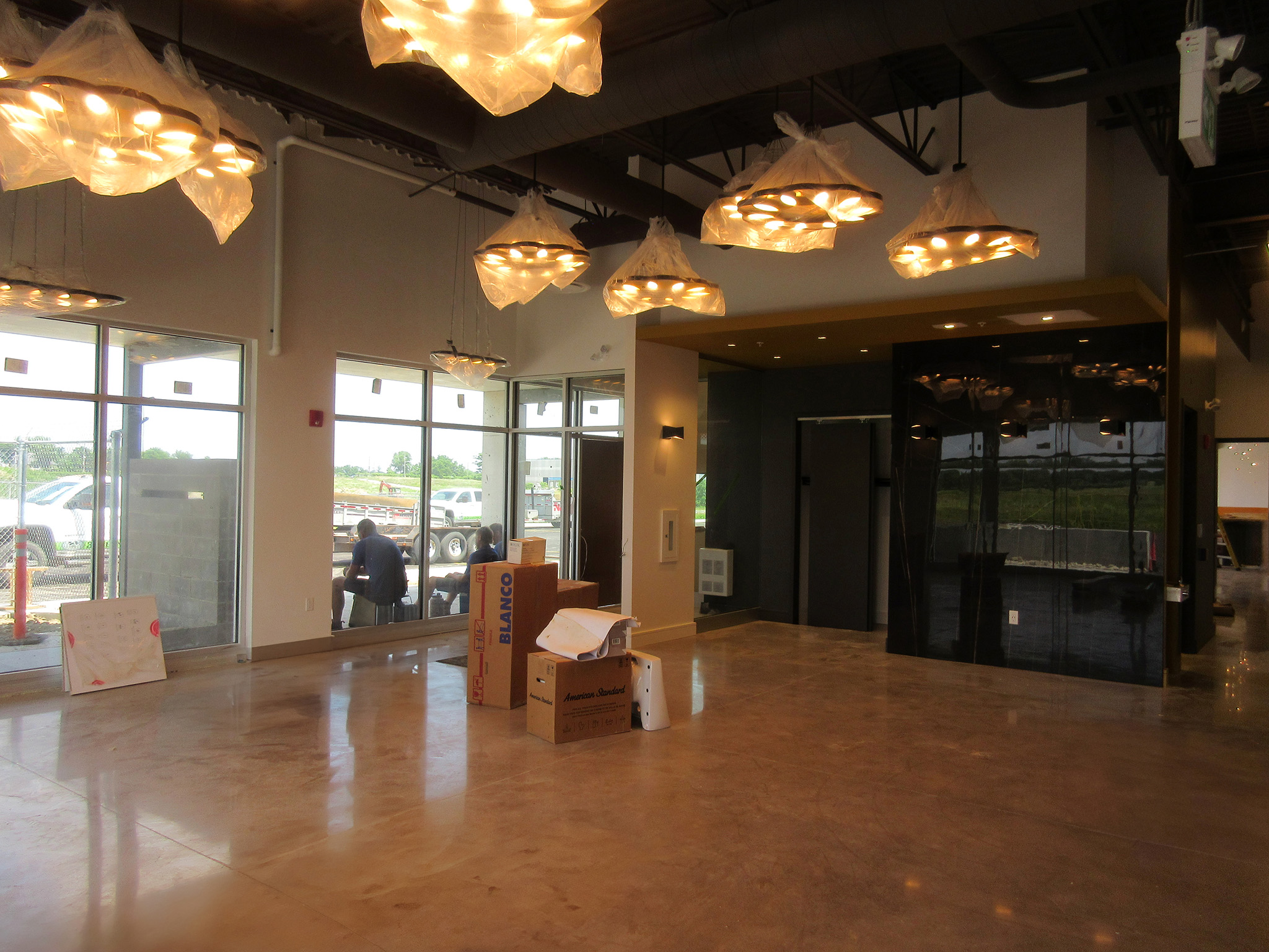 Latitude Air Ambulance 12 Lobby of the Latitude Air Ambulance facility under construction—polished concrete floor, floor‑to‑ceiling windows, sculptural pendant lights wrapped in plastic, and unpacked fixture boxes on the floor.