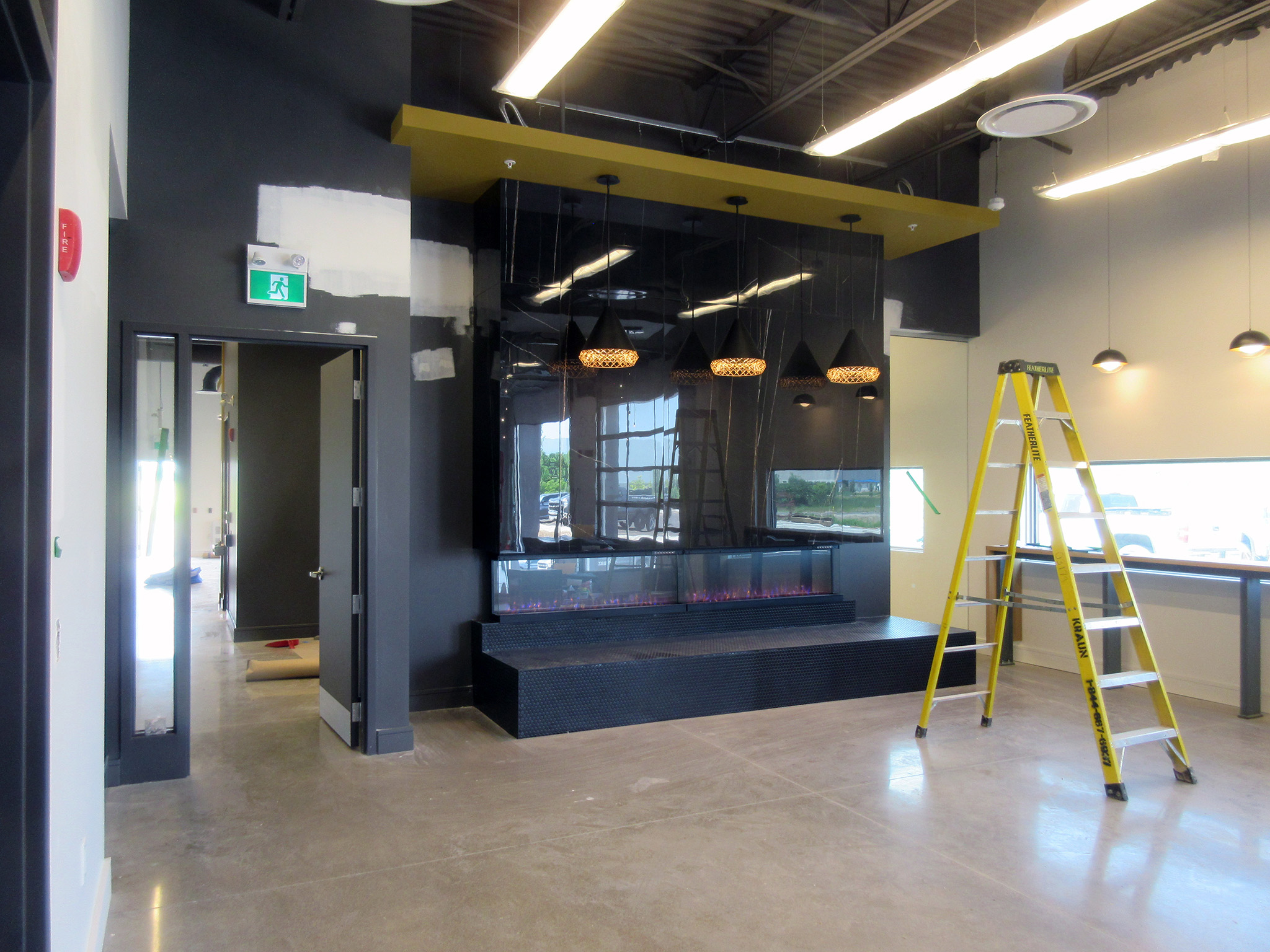 Latitude Air Ambulance 13 Lounge area under construction at the Latitude Air Ambulance facility, showing a glossy black fireplace wall with a linear electric insert, pendant lights hanging from an accent canopy, and a yellow step ladder on the polished concrete floor.