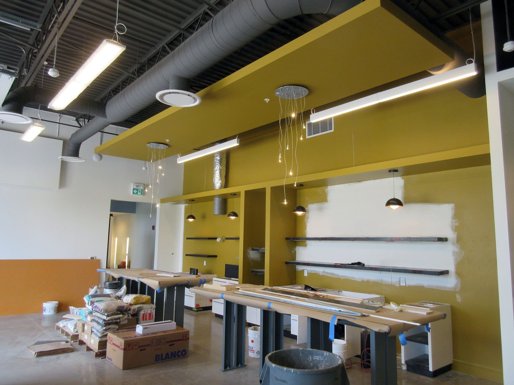 Latitude Air Ambulance 14 Under‑construction staff area at the Latitude Air Ambulance facility with mustard‑yellow accent wall and ceiling panel, exposed grey ductwork, suspended LED and pendant lights, and worktables wrapped in protective paper alongside stacked construction materials.