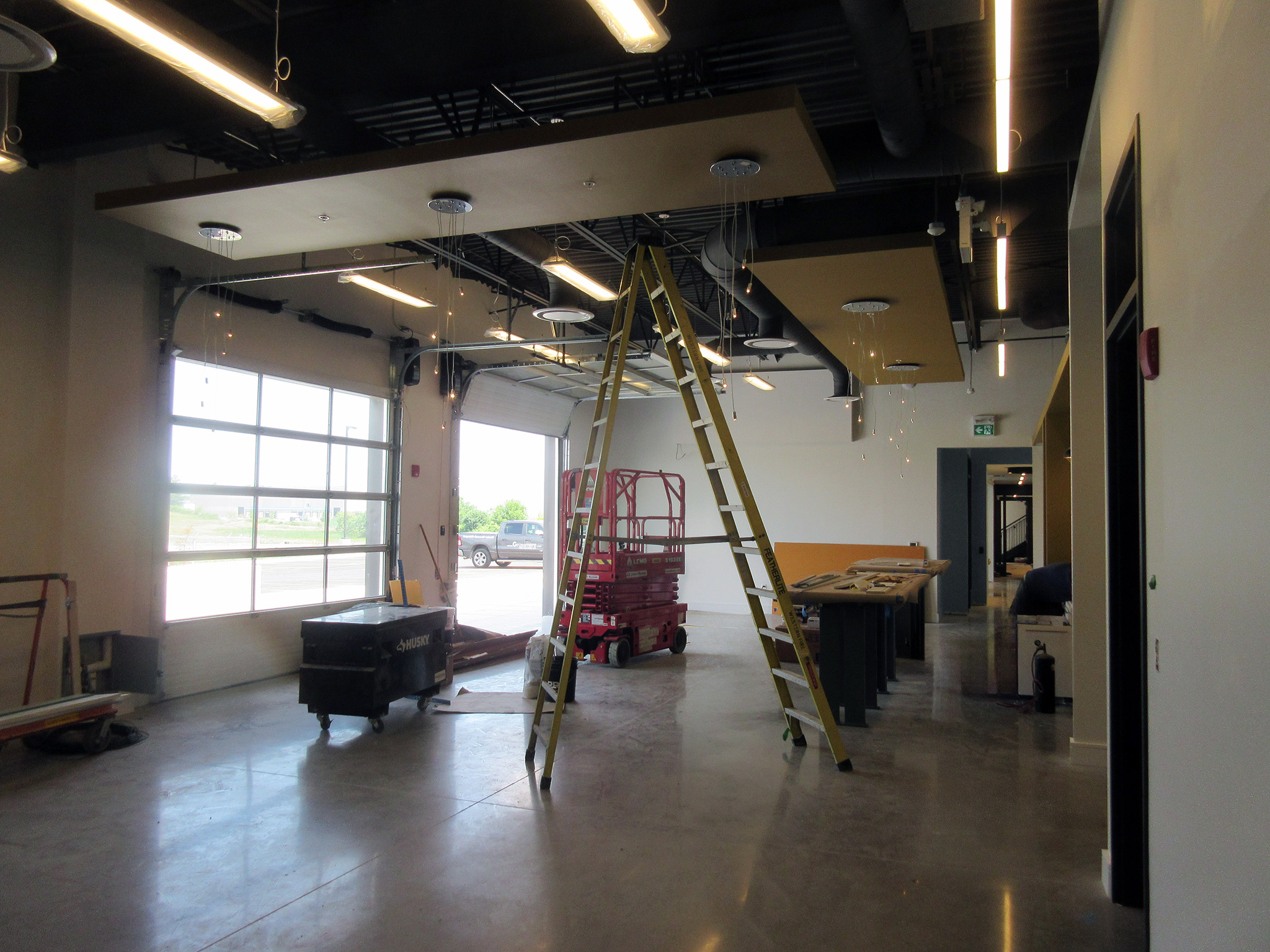 Latitude Air Ambulance 15 Workshop area under construction at the Latitude Air Ambulance facility, featuring suspended ceiling panels, exposed ductwork, a tall yellow ladder, and a red scissor lift near a large glass overhead door.