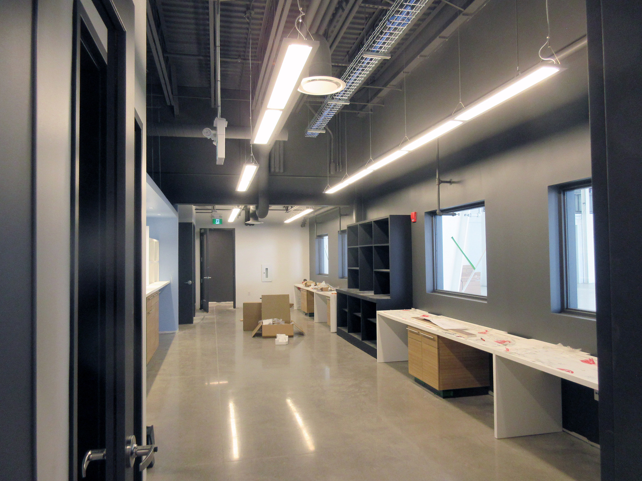 Latitude Air Ambulance 20 Long corridor inside the Latitude Air Ambulance facility with exposed ceiling services and suspended LED strip lights above built‑in work counters, shelving, and windows; cardboard boxes sit on the polished concrete floor during fit‑out.