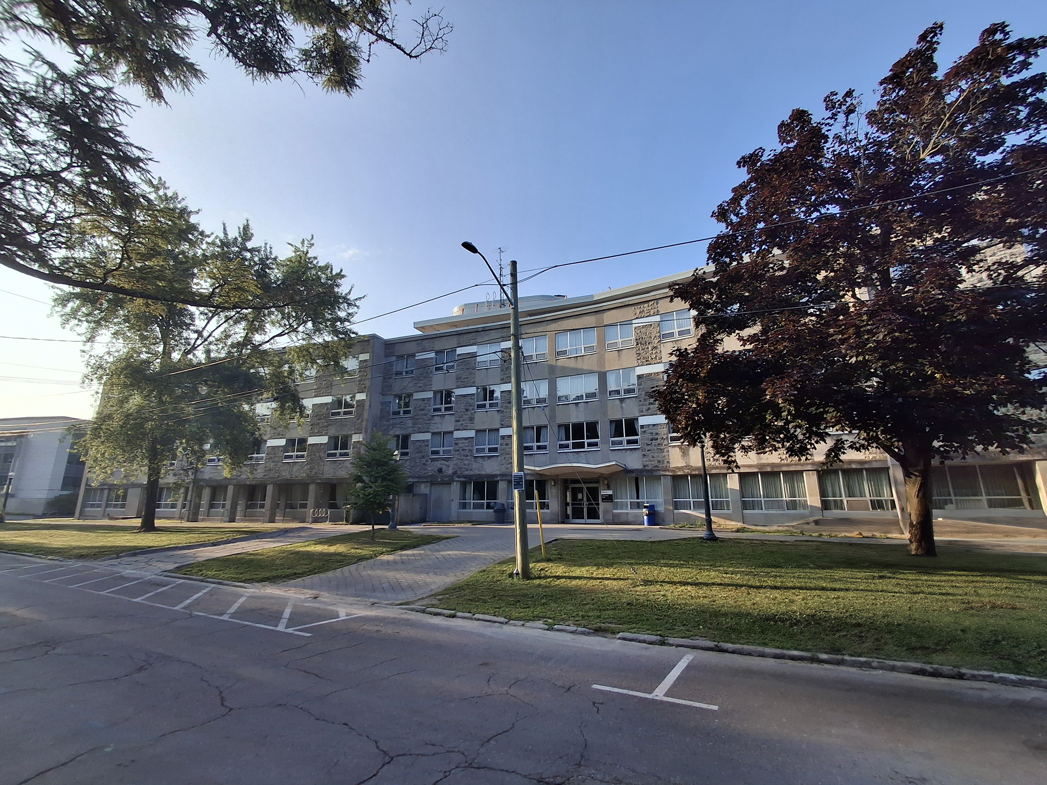 Leonard Hall 11 Street-level wide view of Leonard Hall’s four-storey stone and concrete façade at Queen’s University, framed by trees and a lawn across a quiet campus road.