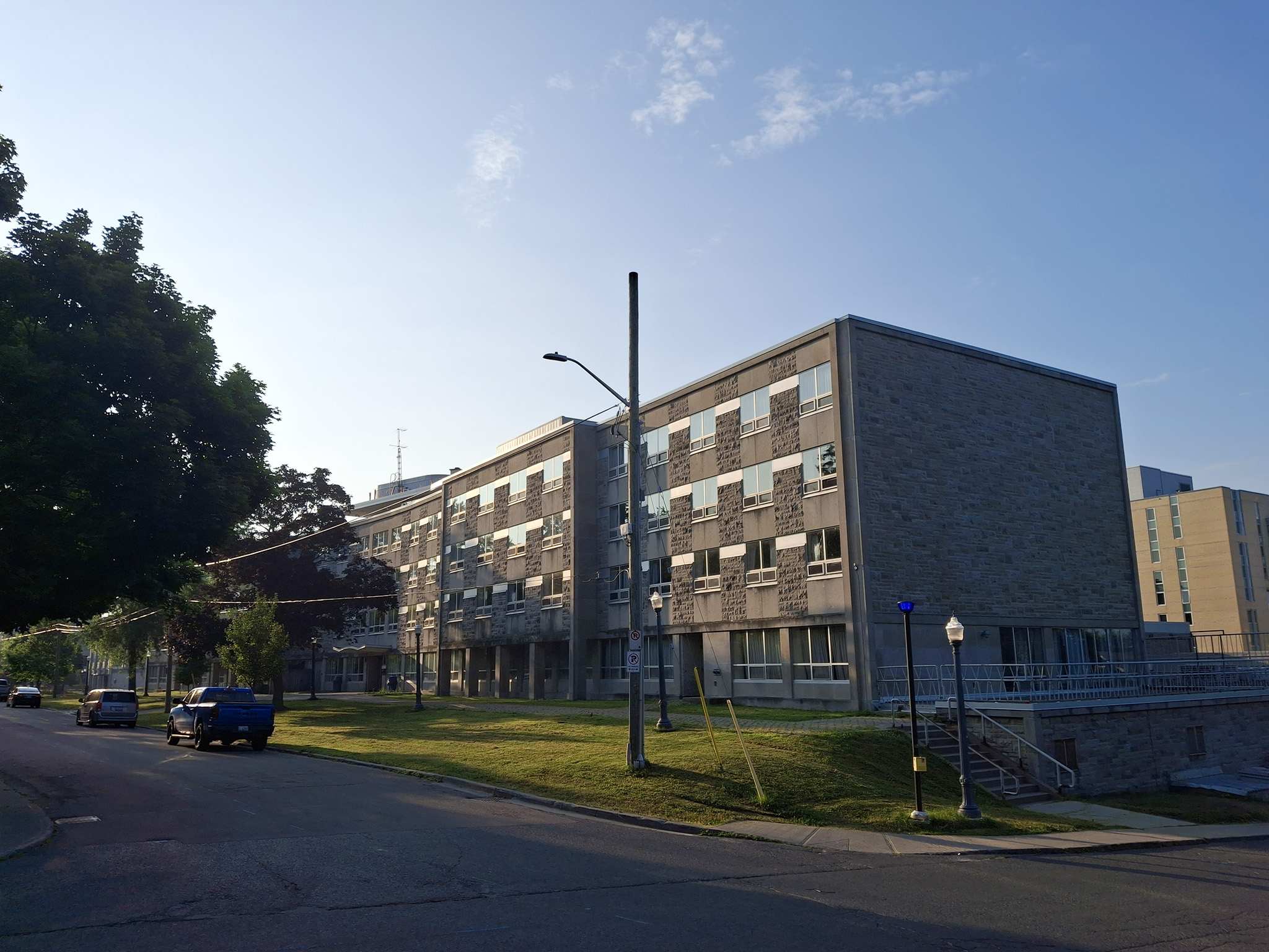 Leonard Hall 12 Angled street view of Leonard Hall’s four-storey stone and concrete façade at Queen’s University, shown in morning light with a lawn, trees, and a few parked cars along the road.