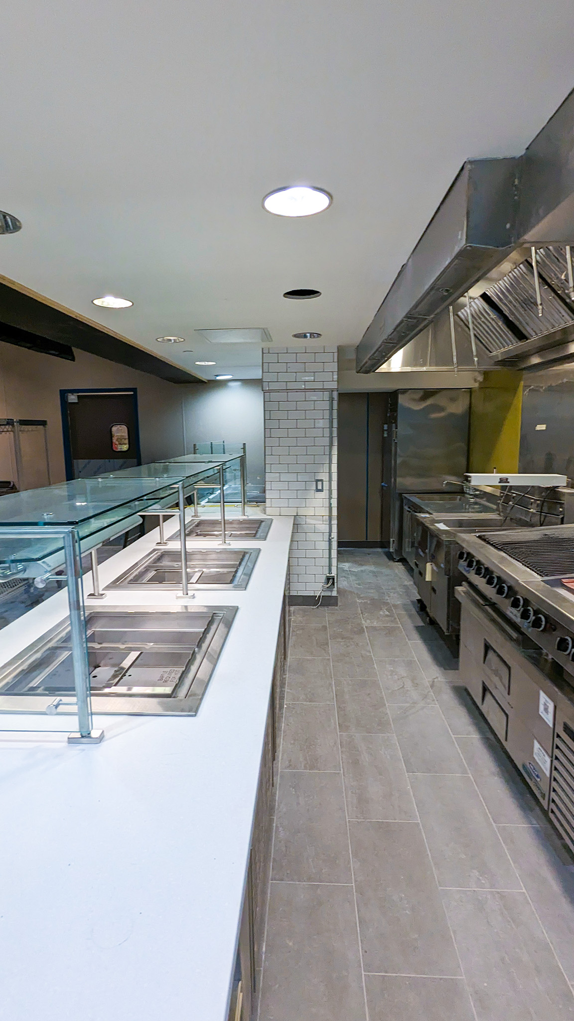 Leonard Hall 2 Interior of a renovated commercial kitchen at Leonard Hall, showing stainless steel cooking equipment, vent hoods, and empty food service stations under glass sneeze guards.