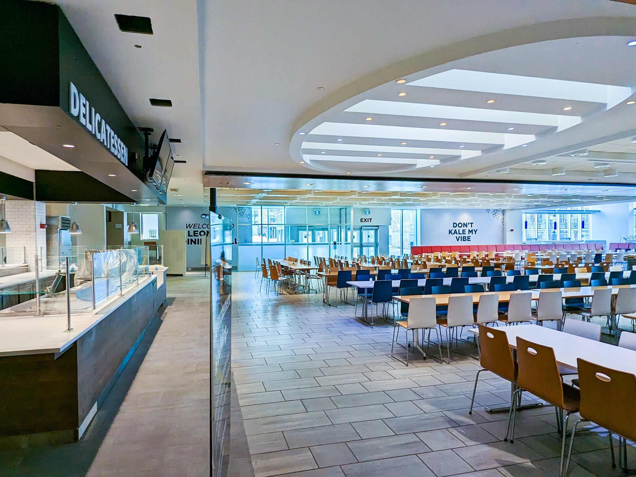 Leonard Hall 5 Wide view of Leonard Hall’s renovated dining hall, with a delicatessen counter on the left and rows of colorful cafeteria tables and chairs under a bright, coffered ceiling; a rear wall reads “DON’T KALE MY VIBE.”