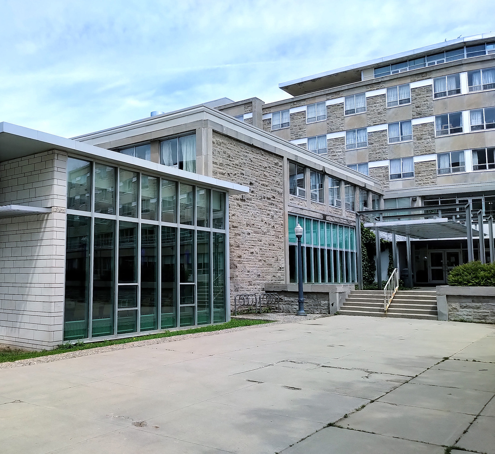 Leonard Hall 7 Side view of Leonard Hall at Queen’s University, showing a modern glass-walled dining hall extension connected to the original stone building, with steps and a covered walkway leading to an entrance.