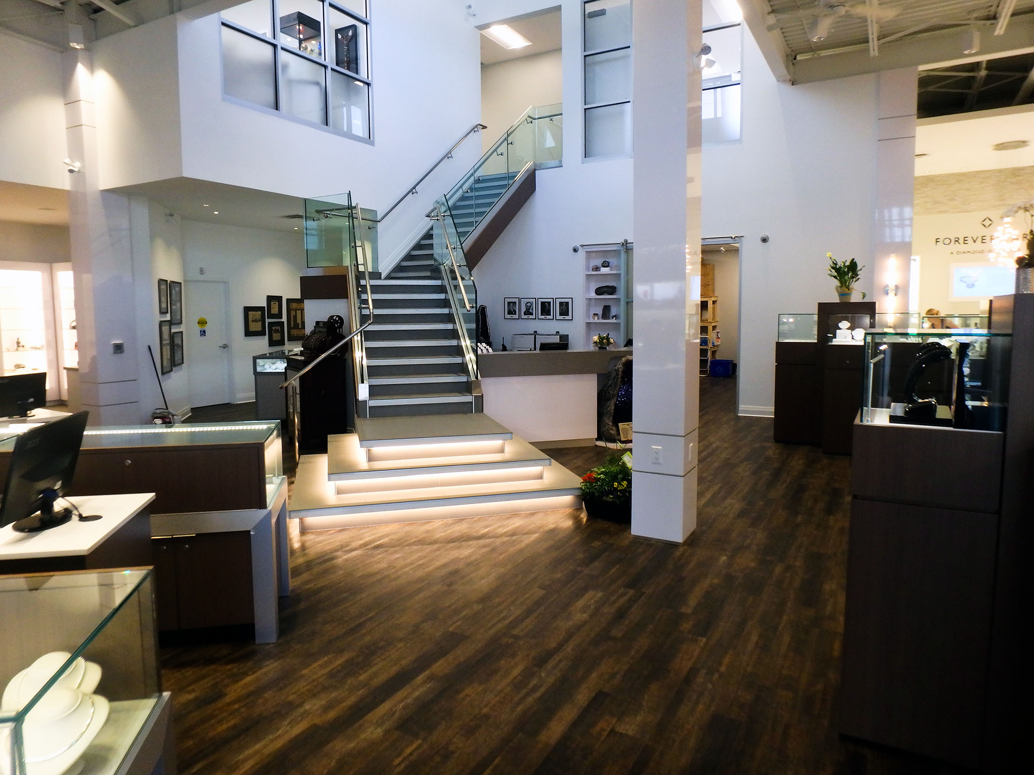 Nash Jewellers 1 Central view of Nash Jewellers showroom featuring a glass-railed staircase with illuminated steps leading to an upper level, surrounded by jewelry display cases and dark wood-grain flooring.