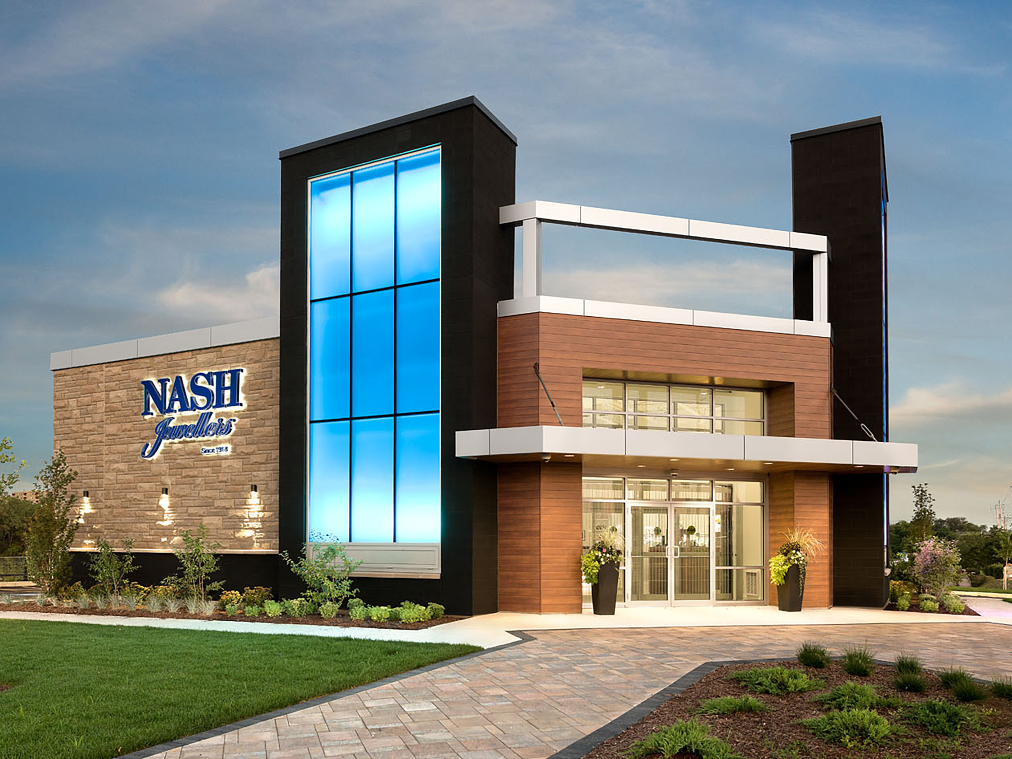 Nash Jewellers 11 Exterior of the Nash Jewellers store in London, Ontario—modern façade with a tall illuminated blue glass tower, stone wall featuring the Nash logo, wood-clad entrance, and landscaped walkway at dusk.