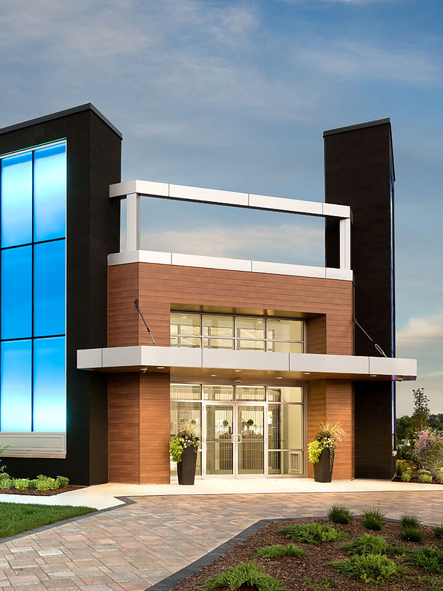 Nash Jewellers 12 Entrance of the Nash Jewellers store featuring a wood-clad façade, glass double doors beneath a white canopy, and an illuminated blue glass tower to the left, with landscaped walkway and planting beds in the foreground at dusk.