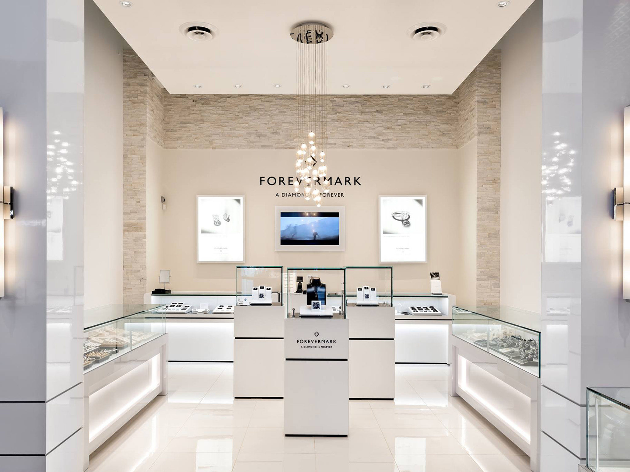 Nash Jewellers 13 Bright Forevermark display area inside Nash Jewellers, featuring glass-topped jewelry cases arranged in a U-shape, illuminated product showcases, and a modern chandelier hanging from a high stone-accent wall.