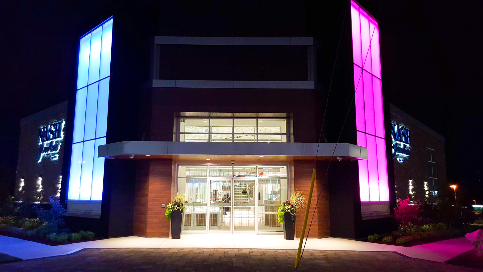 Nash Jewellers 17 Night view of Nash Jewellers’ storefront with blue-lit glass tower on the left and pink-lit tower on the right, framing the brightly illuminated main entrance.