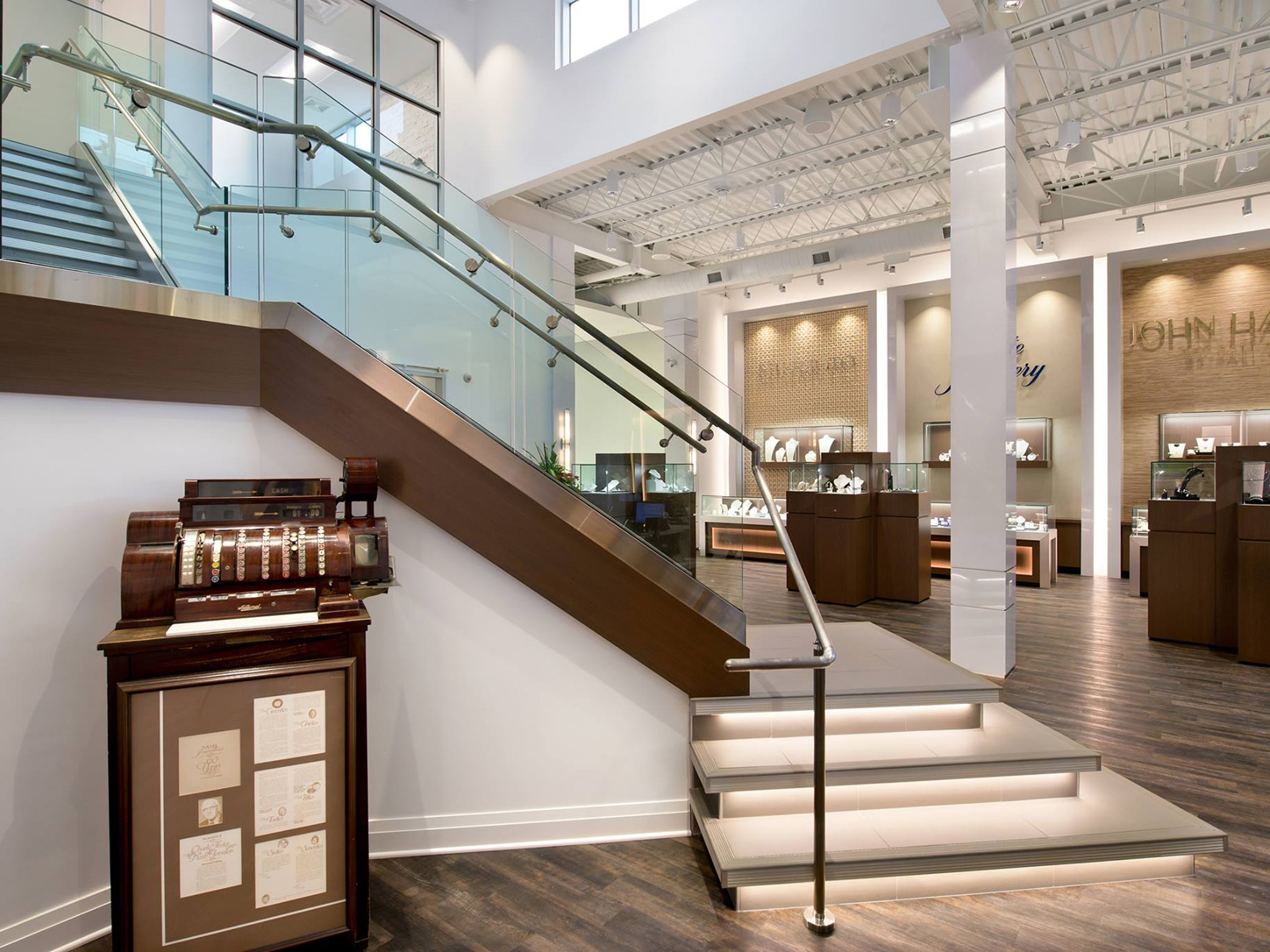 Nash Jewellers 18 Interior of Nash Jewellers showroom showing a glass-railed staircase with wood accents, under-lit steps, a vintage wooden cash register display, and illuminated jewelry cases beneath an exposed white ceiling.