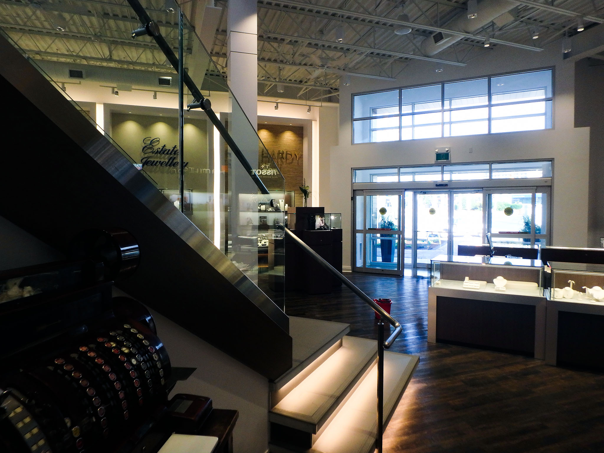Nash Jewellers 4 Interior view toward Nash Jewellers’ glazed entrance doors, with an under-lit glass-railed staircase in the foreground and jewelry display cases on dark wood flooring.