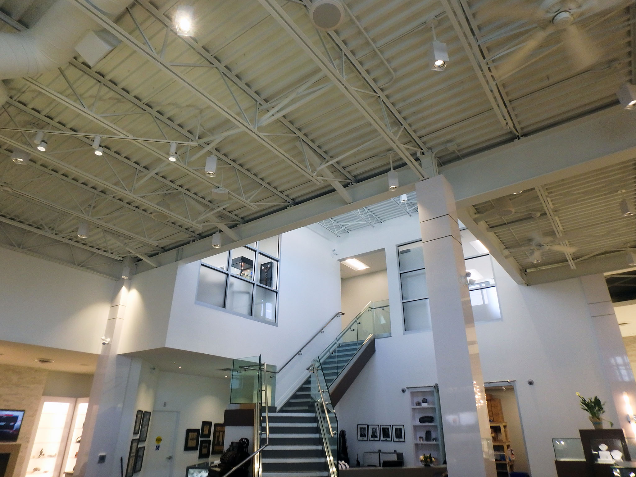 Nash Jewellers 5 Upward view inside Nash Jewellers showing the exposed white structural ceiling with track lighting and a central glass-railed staircase leading to a mezzanine level.
