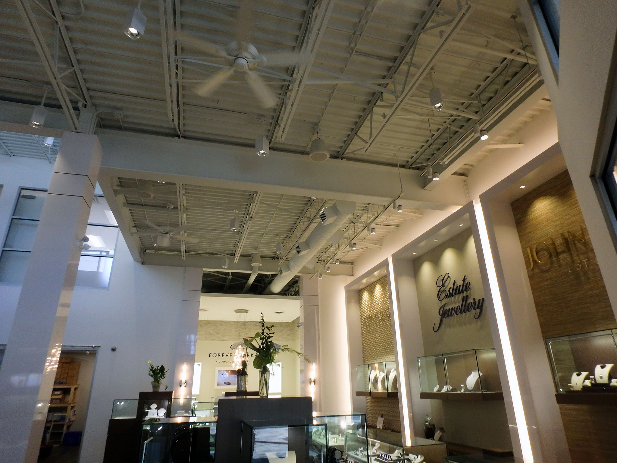 Nash Jewellers 6 View across Nash Jewellers showroom highlighting the exposed white structural ceiling with track lights and fans, and illuminated “Estate Jewellery” wall niches displaying jewelry beneath wood-textured panels.
