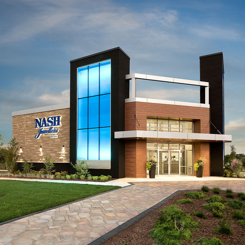Exterior of the Nash Jewellers store in London, Ontario—modern façade with a tall illuminated blue glass tower, stone wall featuring the Nash logo, wood-clad entrance, and landscaped walkway at dusk.