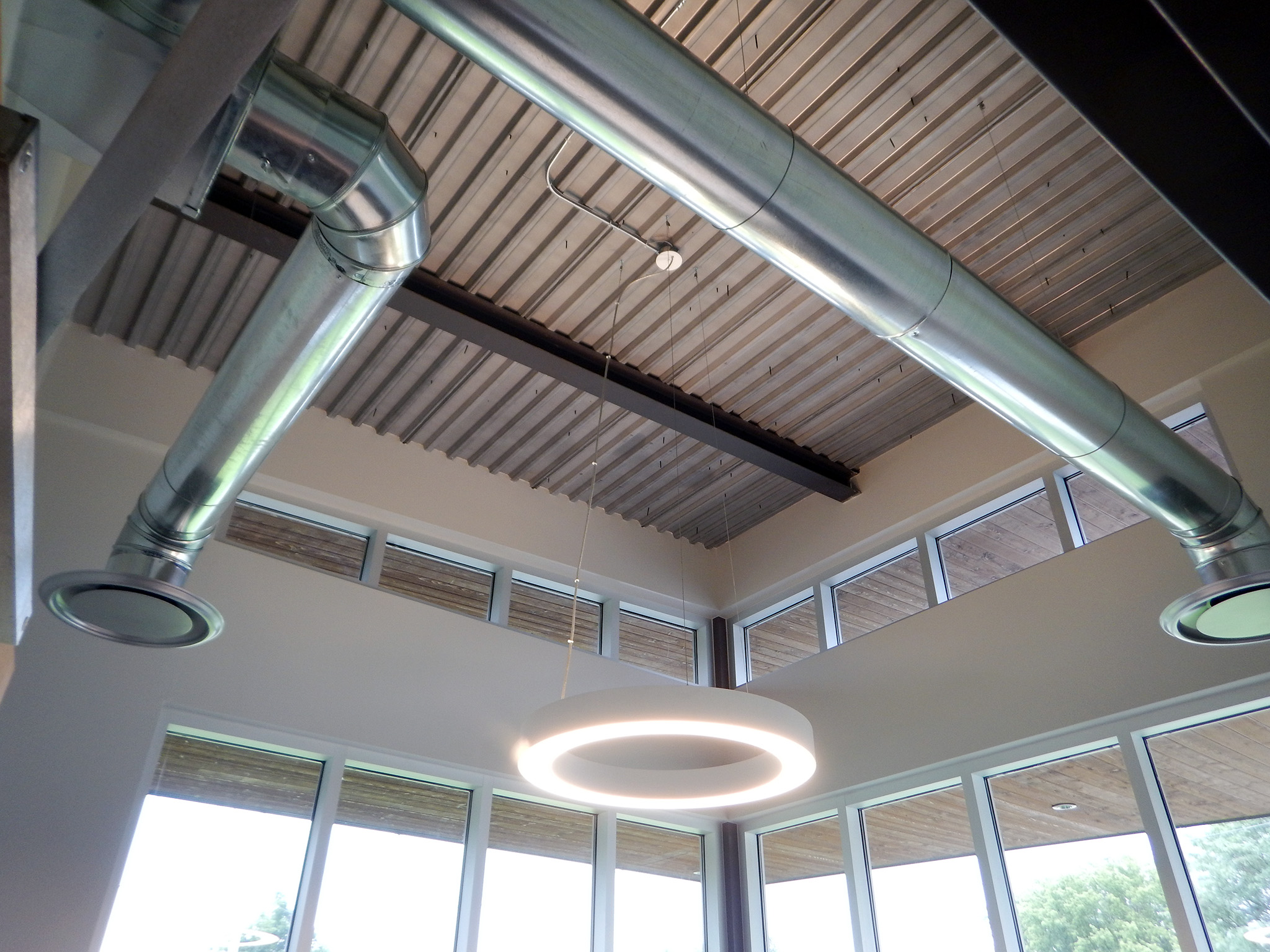L'Arche 1 Interior view of exposed metal ductwork and circular LED light fixture against a high windowed wall.