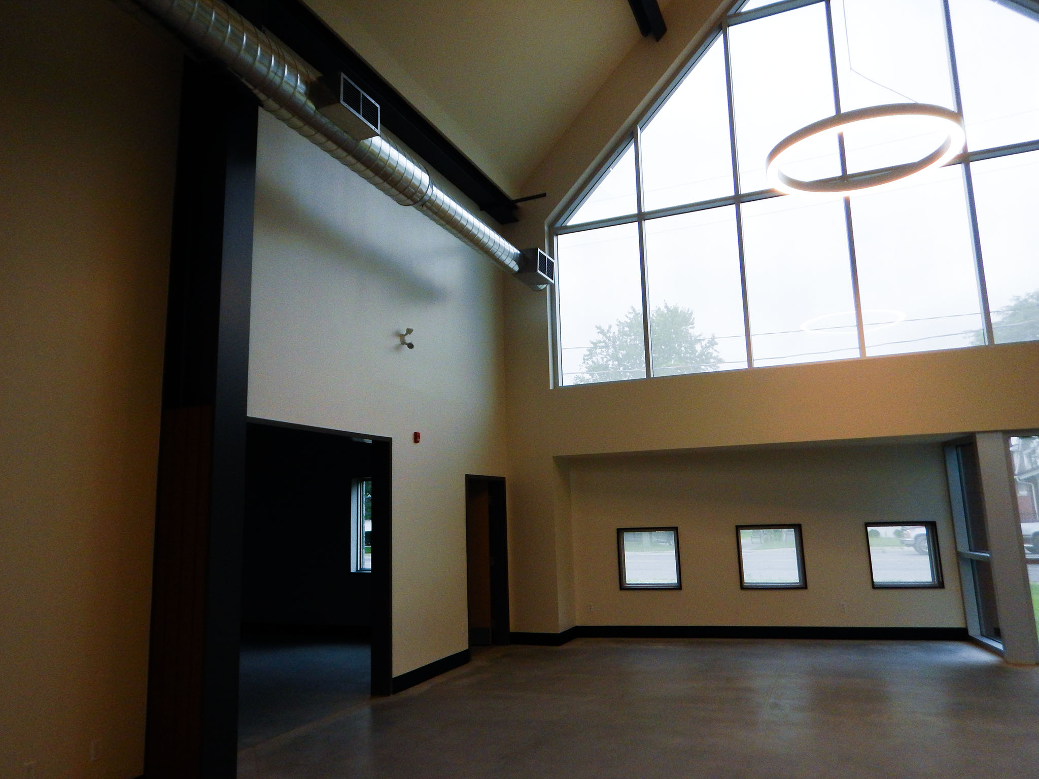 L'Arche 6 Great Hall interior at the L’Arche Gathering Facility with tall windows, circular LED light, and exposed ductwork.