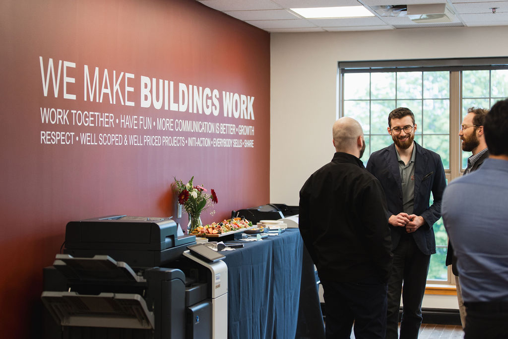 Group of people talking in office near wall with 'We Make Buildings Work' slogan.