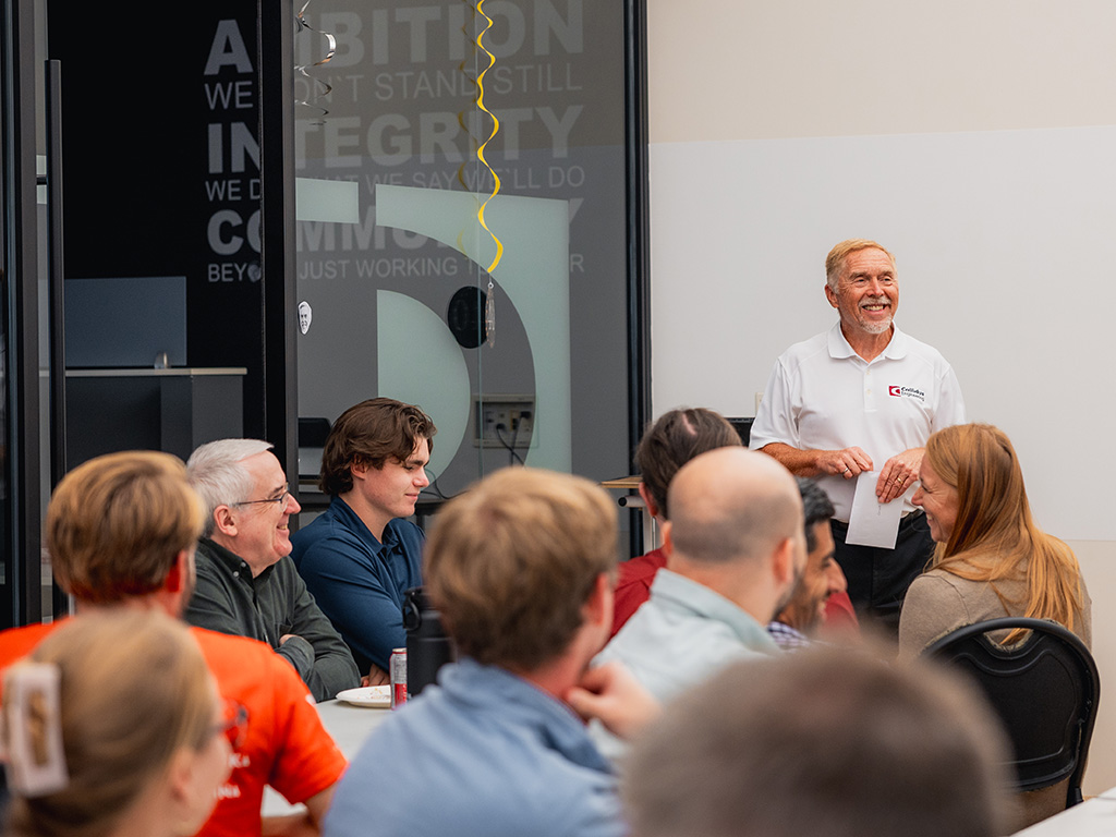 Grant Hall 1 Grant Hall, wearing a white Callidus Engineering polo shirt, smiles while standing and holding a paper in front of a seated group of colleagues during a gathering. Staff members face him around a table, and motivational words such as “Ambition,” “Integrity,” and “Community” are visible on the glass wall behind.
