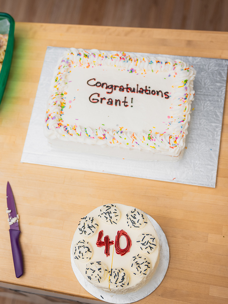 Grant Hall 2 Two celebration cakes on a wooden table. The larger rectangular cake with rainbow sprinkles reads “Congratulations Grant!” in red icing, and the smaller round cake is decorated with black sprinkles and the number “40” in red icing. A purple knife rests beside the cakes.