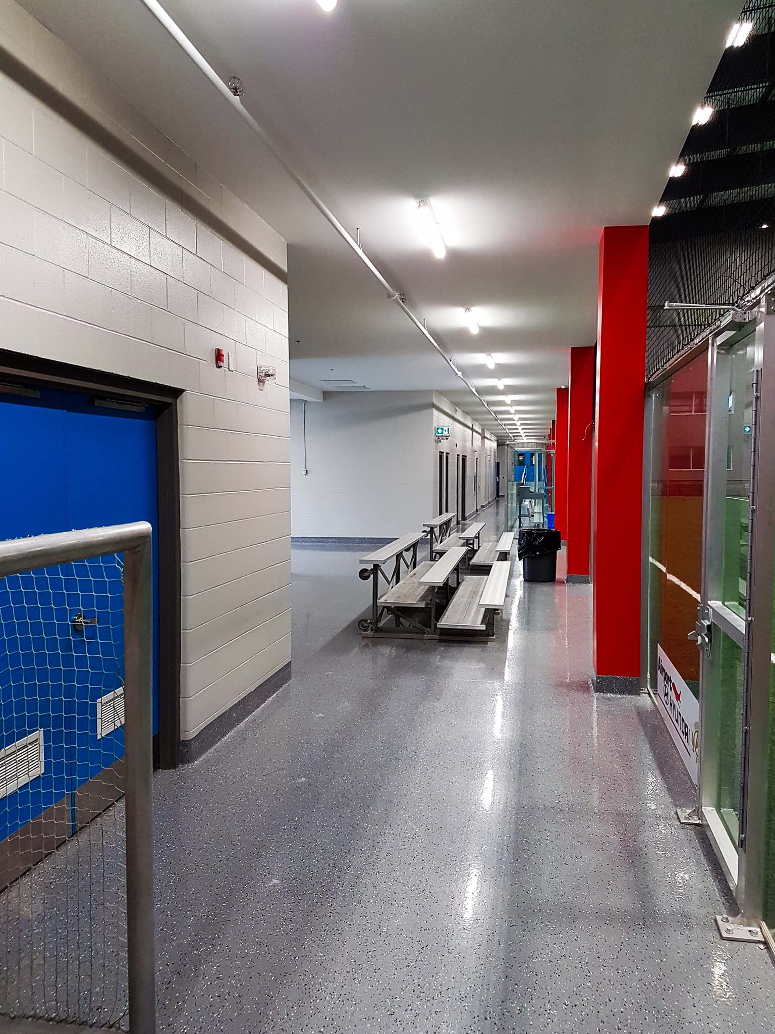 BMO Centre (10) Indoor corridor with polished floors, red columns, metal bleachers, and glass partitions overlooking the turf field.