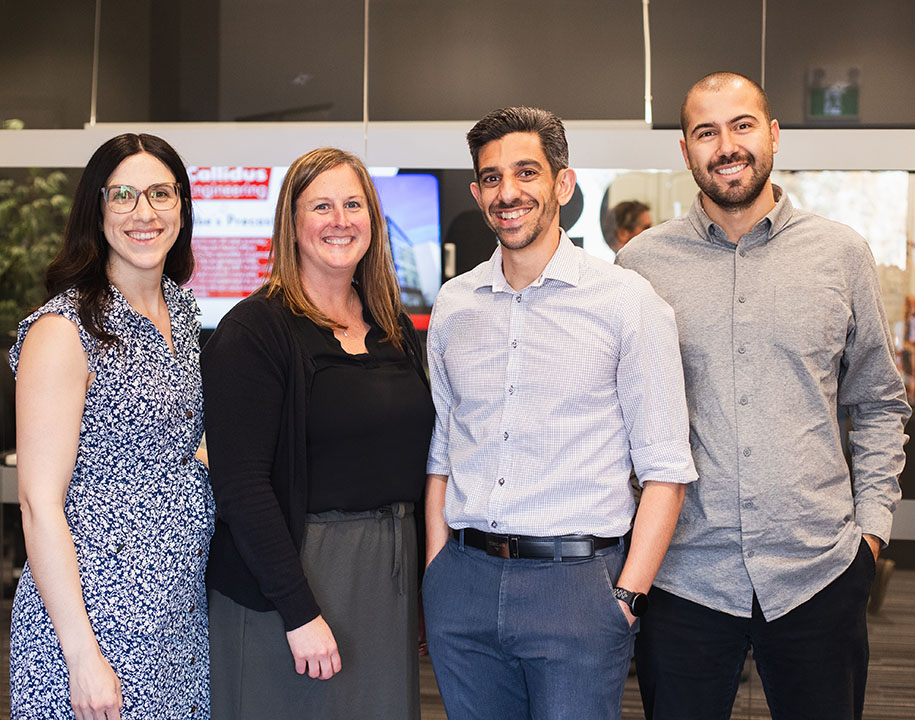 Four Callidus Engineering team members stand together smiling in an office setting. The group includes two women and two men in professional attire, with glass walls, plants, and a digital display visible in the background.