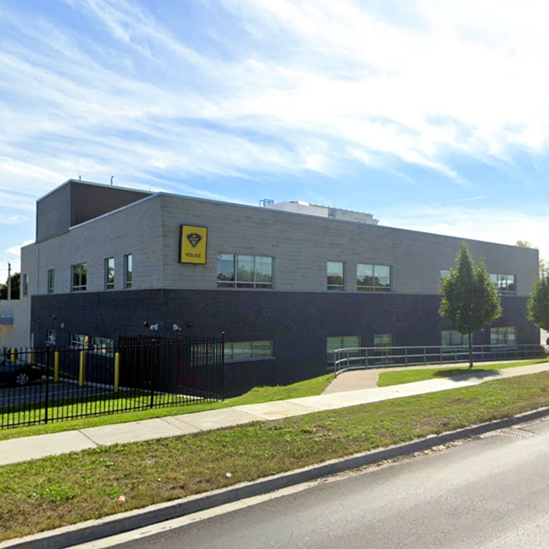Quinte West OPP Exterior view of the Quinte West OPP Detachment, showing a two-storey grey and dark brick building with accessible walkways, small trees along the sidewalk, and a police sign on the upper level.