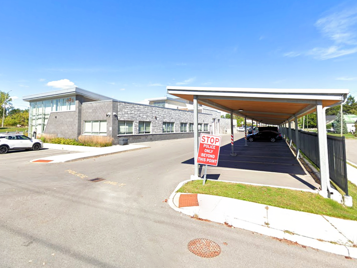 Exterior view of the Quinte West OPP Detachment showing the building’s front elevation, adjacent parking area, and a covered parking structure with a sign that reads “Stop, Police Only Beyond This Point.