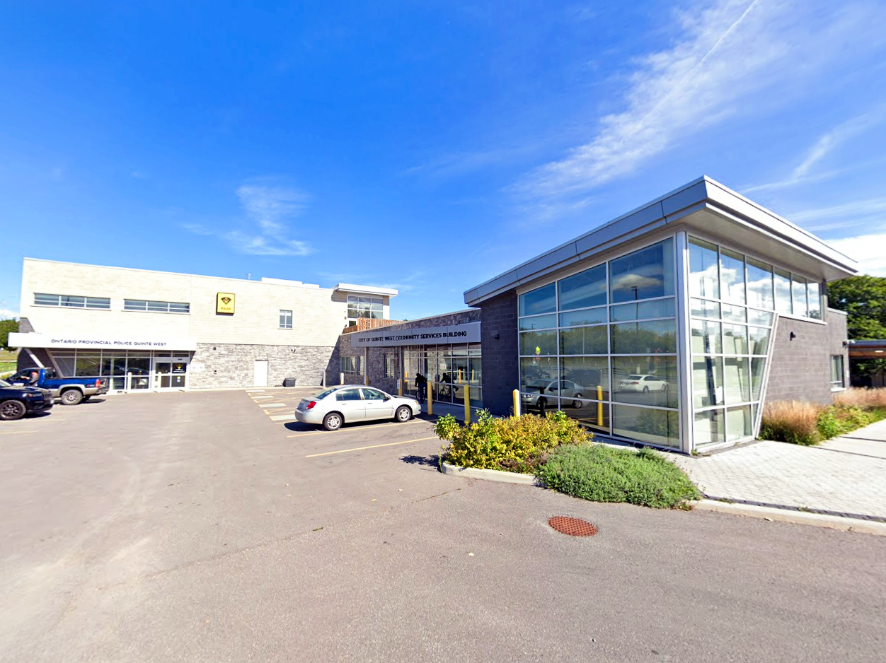 Front view of the Quinte West OPP Detachment and the adjacent Community Services Building, showing large glass windows, parking spaces, and the main entrances to both buildings.