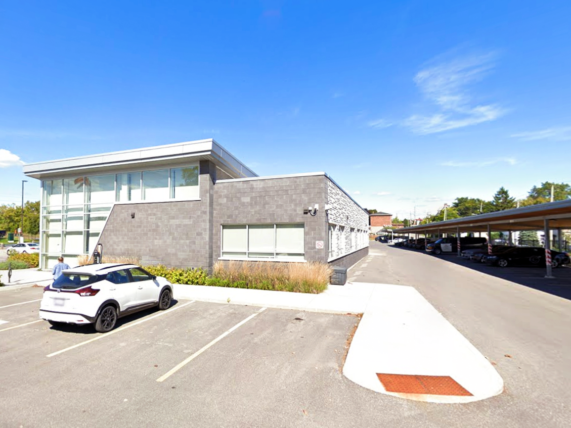 Side view of the Quinte West OPP Detachment and Community Services Building, showing parked vehicles, landscaping, and the covered parking structure extending along the driveway.