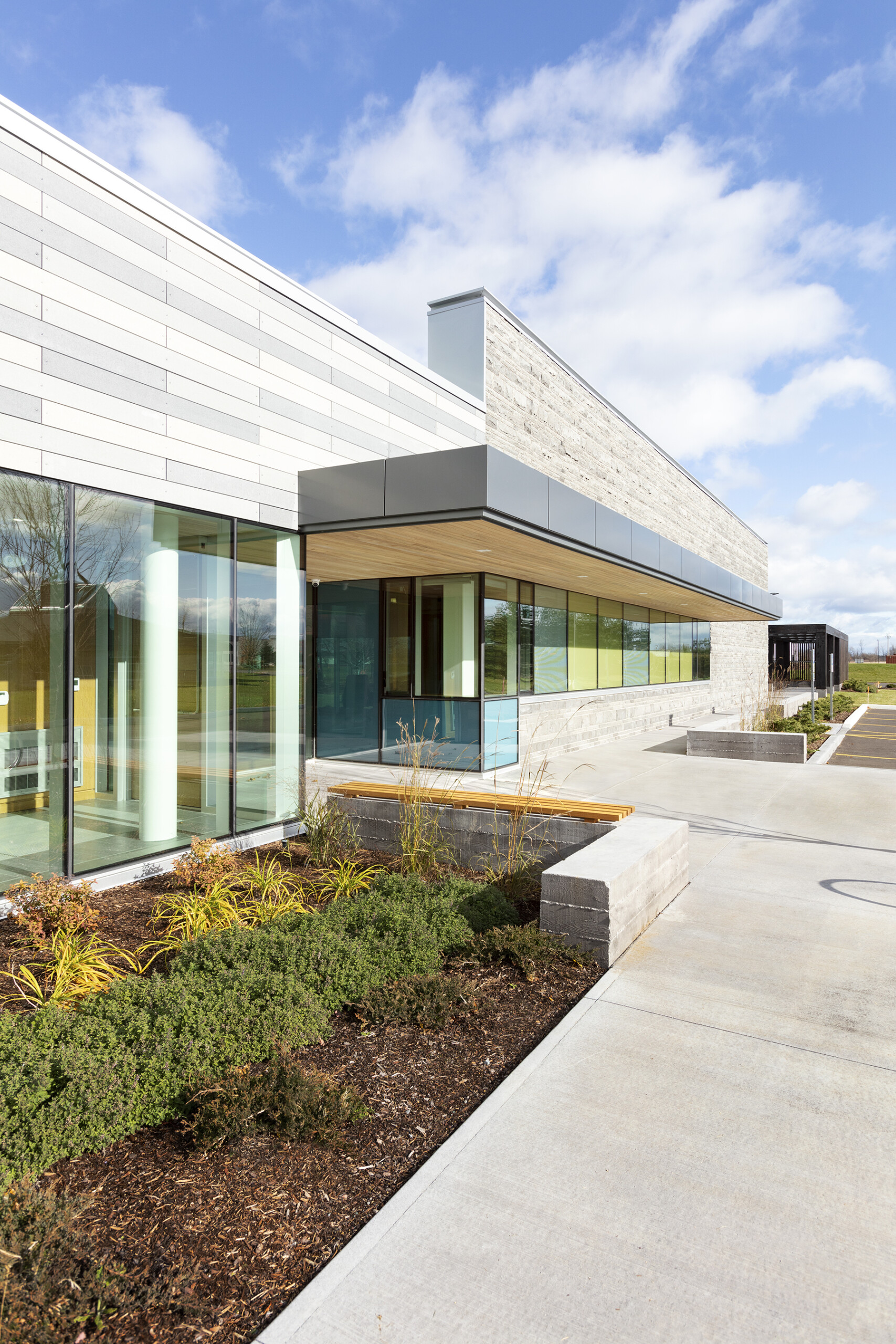 © krista jahnke photography Exterior of a modern single-storey building with glass walls, light stone cladding, landscaped plantings, and a concrete walkway under a blue sky.