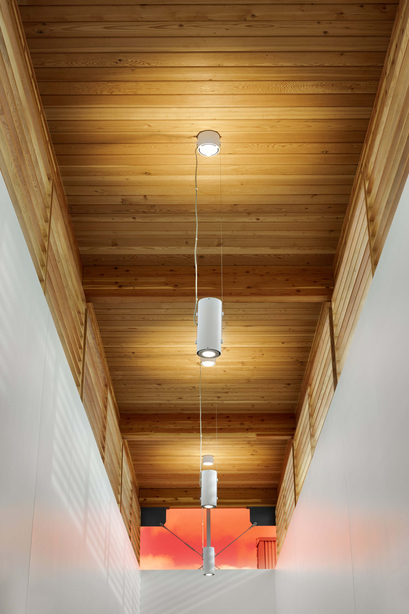 © krista jahnke photography Interior view of a corridor with a wood ceiling, hanging cylindrical light fixtures, and white walls with warm reflected light.