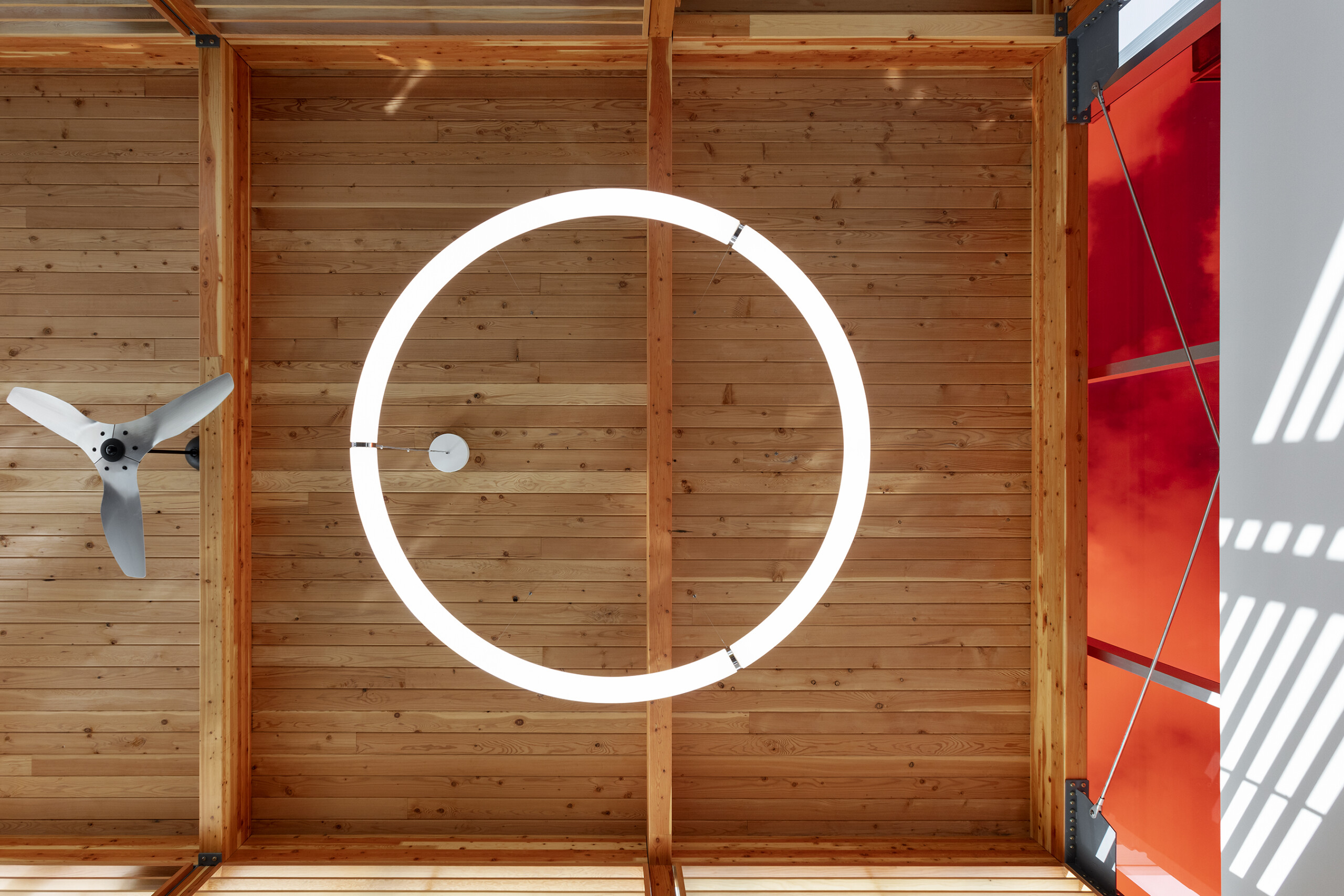 © krista jahnke photography Wood ceiling with exposed beams, a large circular light fixture, and a ceiling fan beside a red wall panel.