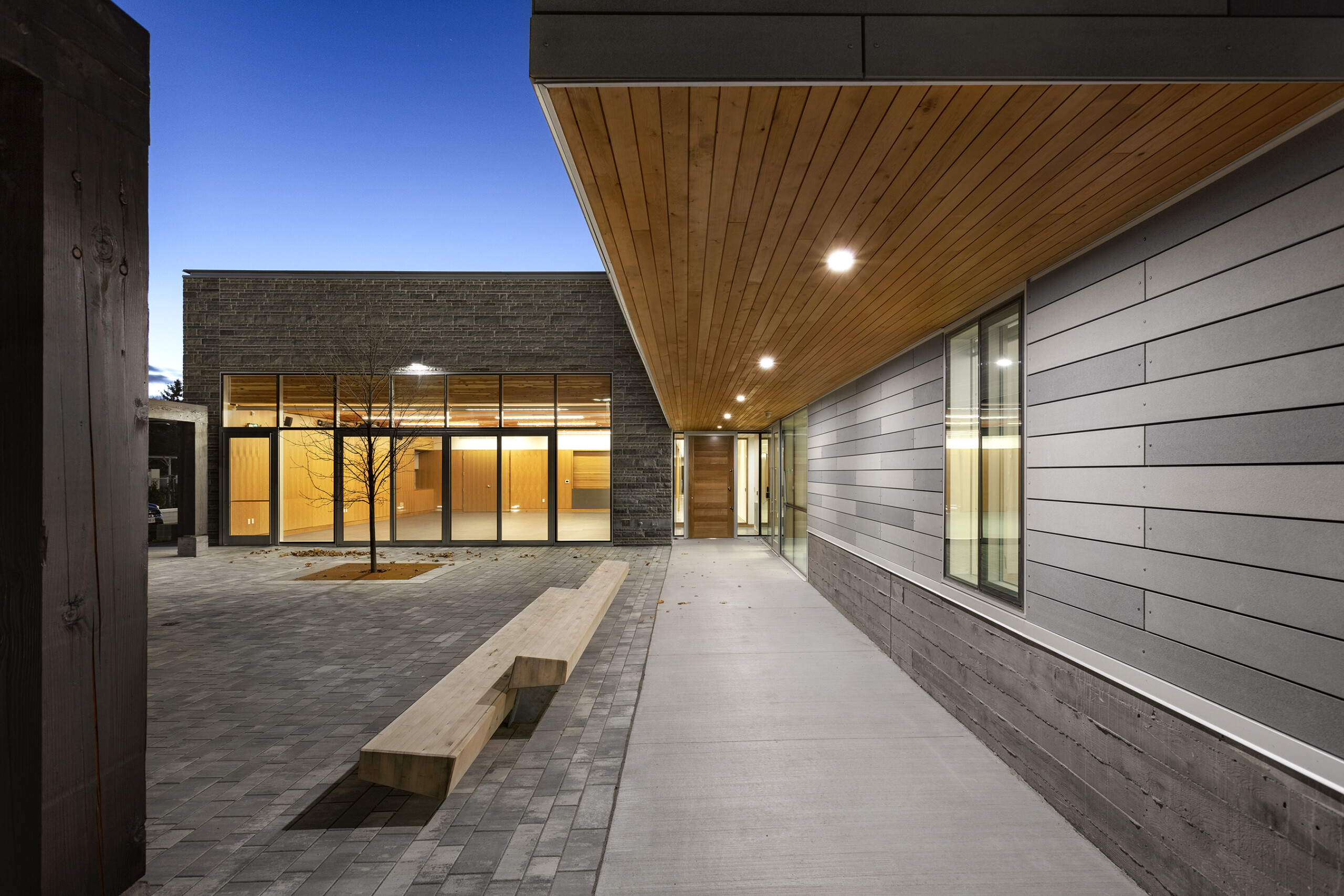 © krista jahnke photography Evening view of an exterior courtyard with paving, a wooden bench, glass walls, and a wood-clad overhang with recessed lights.
