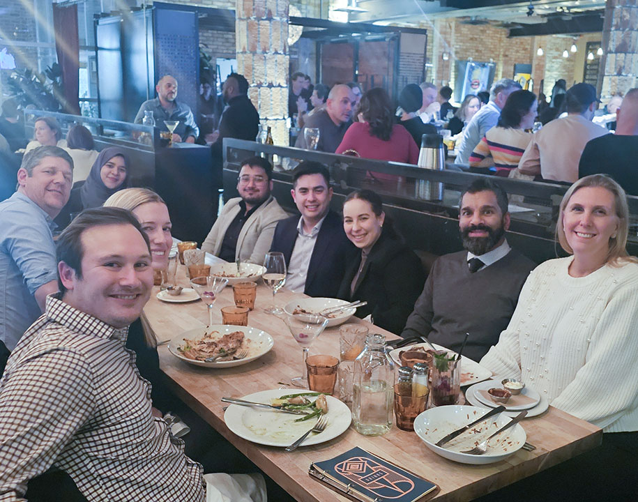Group of Callidus Engineering staff seated together at a long dining table in a restaurant, smiling toward the camera during the Kitchener-Waterloo office holiday party.