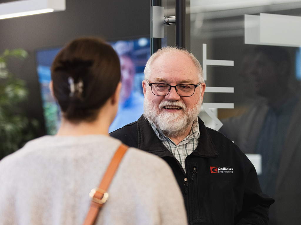 Older man wearing glasses and a black Callidus Engineering jacket speaks with another person in a modern office setting, smiling during a casual conversation near a glass wall.