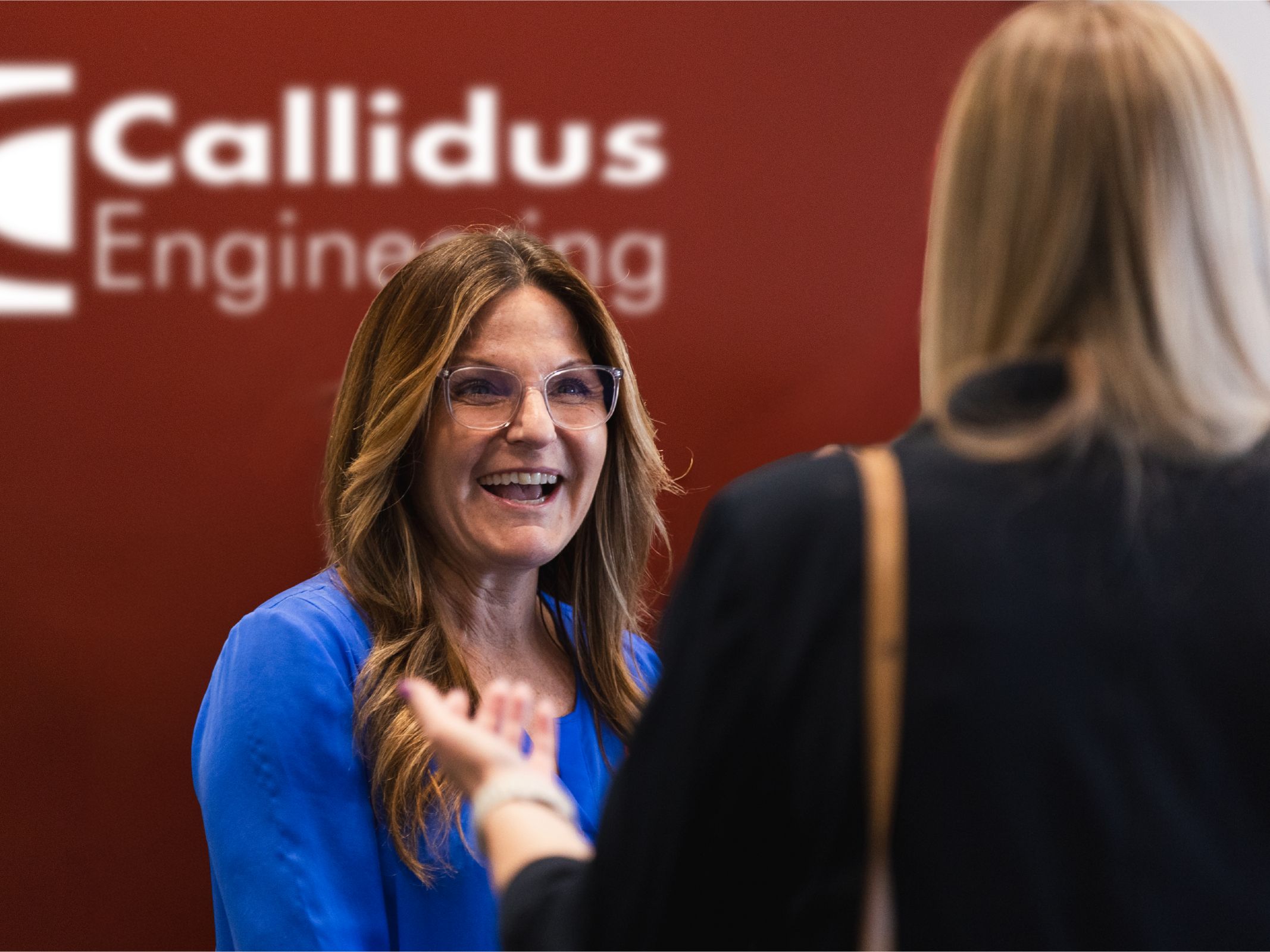 Two people talking in a Callidus Engineering office reception area. A woman in a blue blouse and glasses smiles while speaking to another person seen from behind. The Callidus Engineering logo is visible on a red wall behind them.