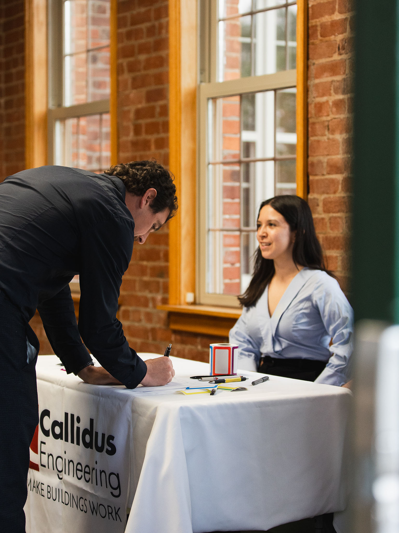 A person signs in at a table covered with a Callidus Engineering tablecloth while speaking with a seated staff member in a light blue blouse. The table is set up inside a brick-walled space with large windows.