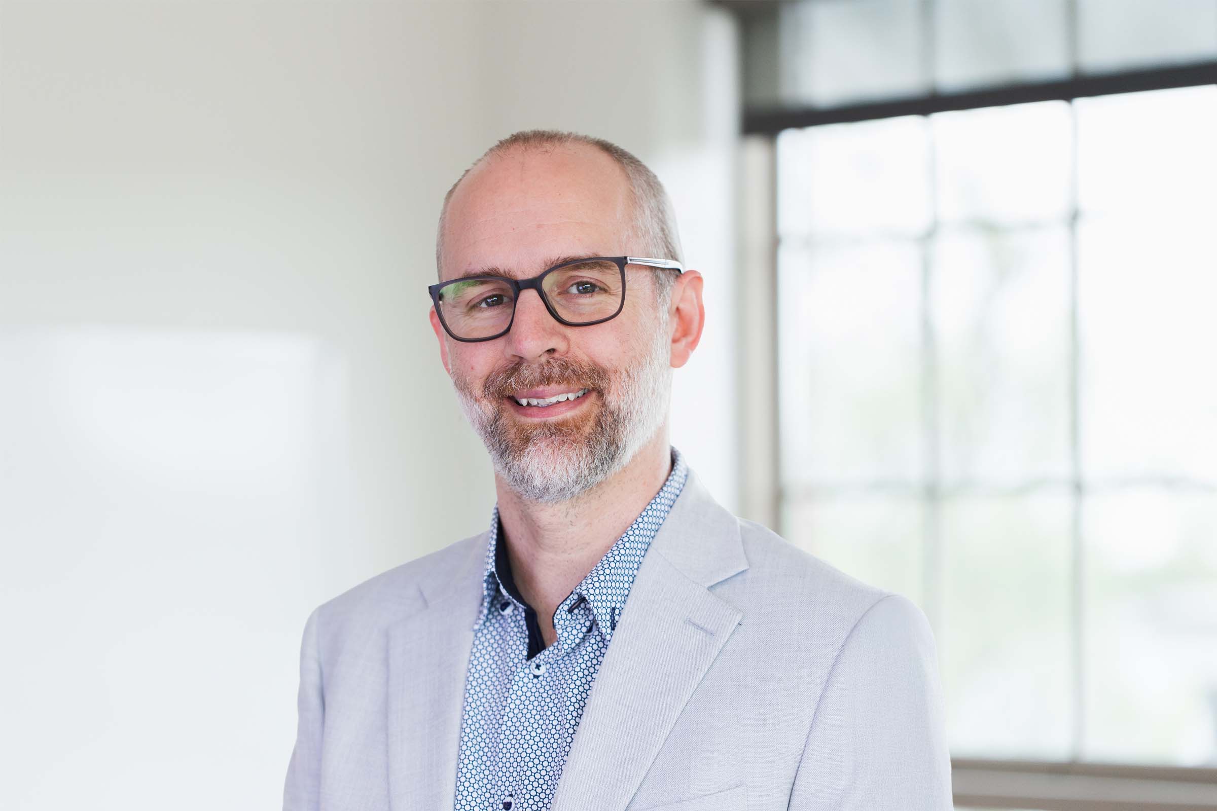 Derek Reid Professional headshot of a smiling man wearing glasses and a light blazer in a bright office setting.
