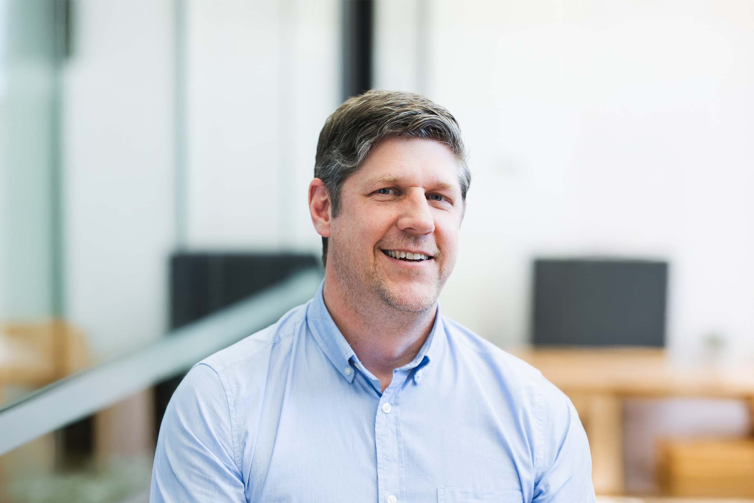 Professional headshot of a smiling man in a light blue shirt seated in a bright office setting.