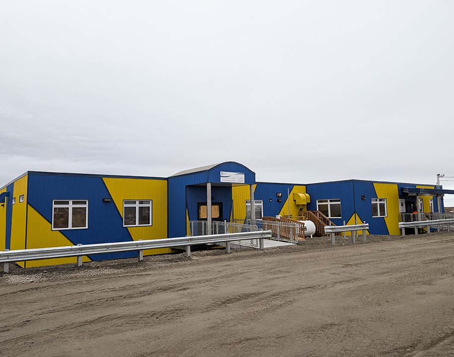 Blue and yellow Nattiqsuqtiit Multi-Use Facility beside a gravel road in Clyde River, Nunavut.