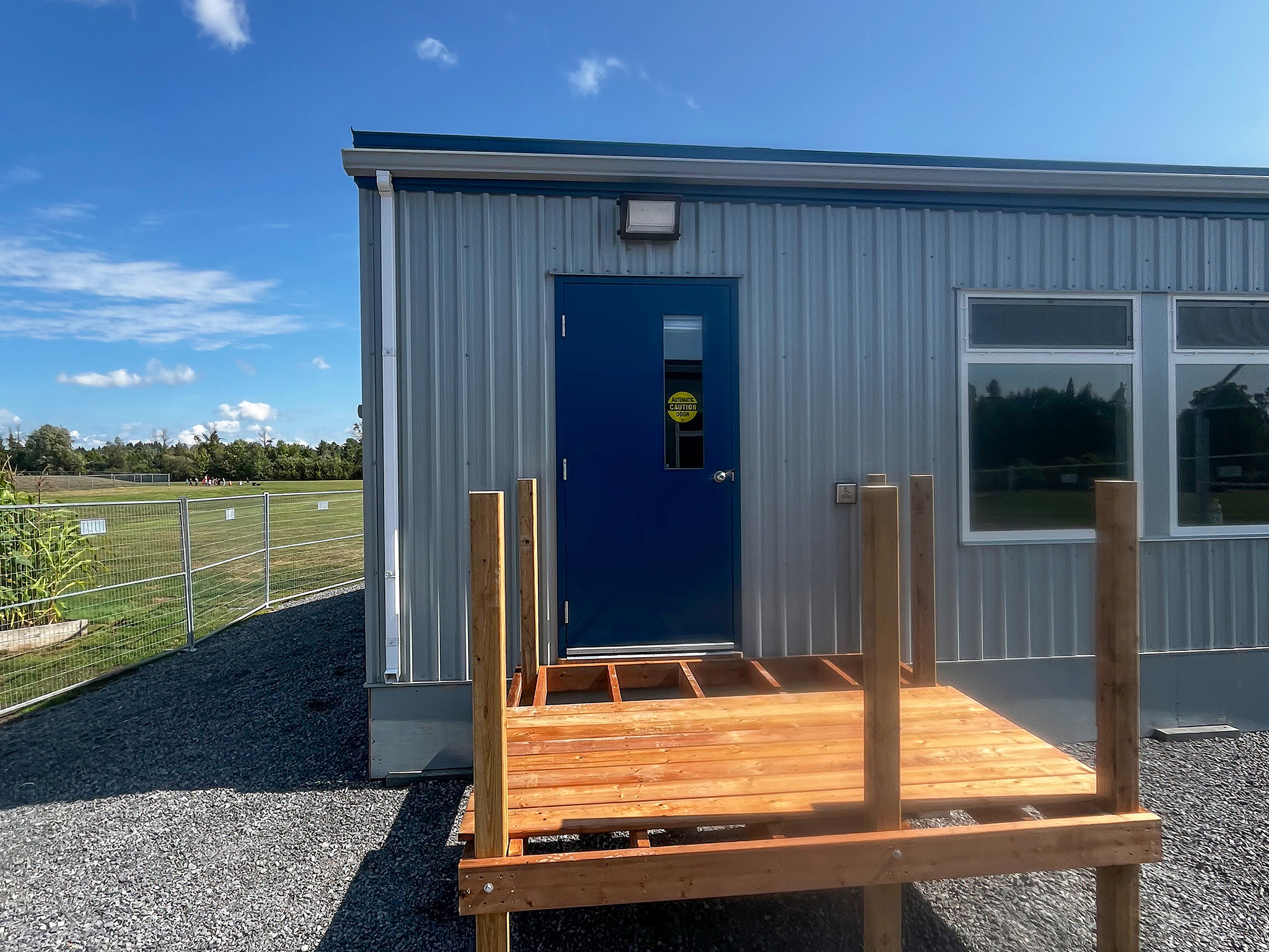 Beckwith Public School (1) Grey portable classroom with a blue entrance door, wooden platform, and playing field visible beyond.