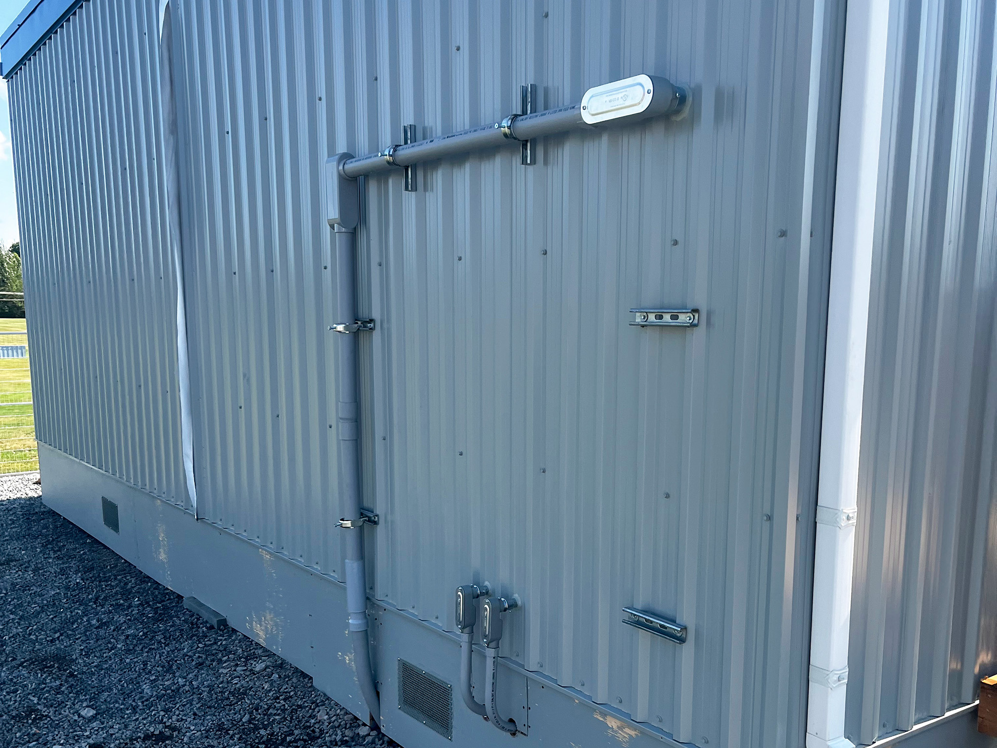 Beckwith Public School (4) Exterior wall of a portable classroom with grey metal siding, surface-mounted conduits, electrical boxes, and a ventilation grille.