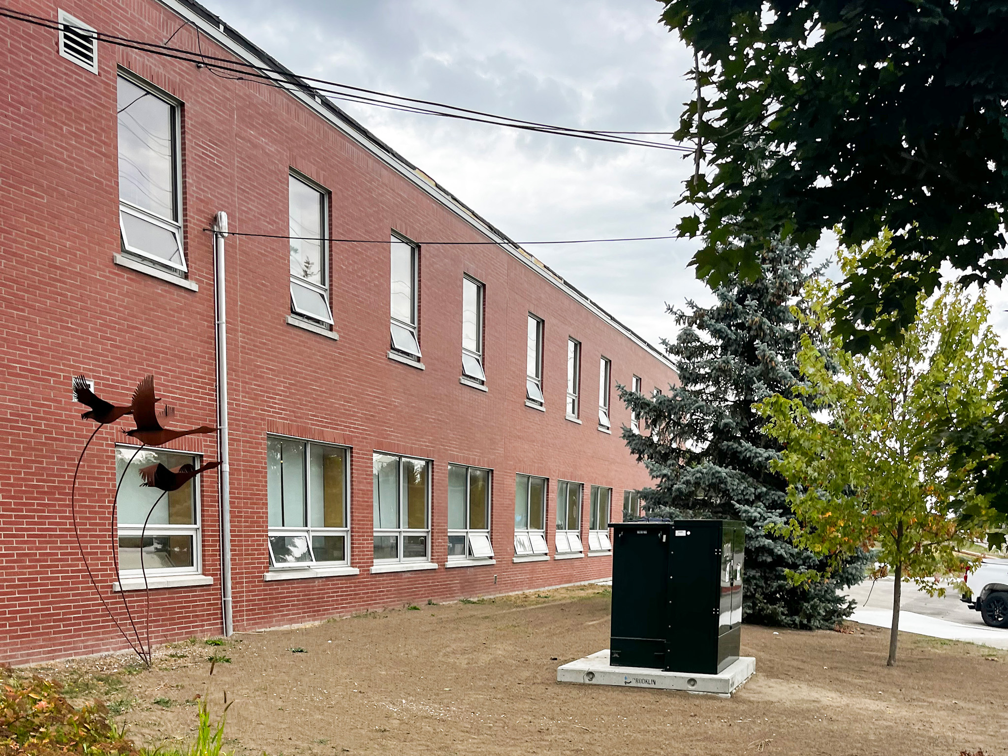 Elginburg Public School (1) Exterior of a two-storey brick school building with a large black utility cabinet, trees, and overhead power lines.