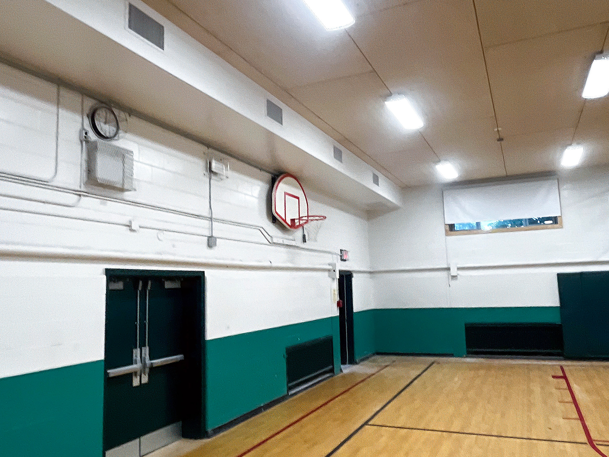 Elginburg Public School (3) School gymnasium with wall-mounted ventilation grilles, ceiling lights, a basketball hoop, and green-and-white walls.